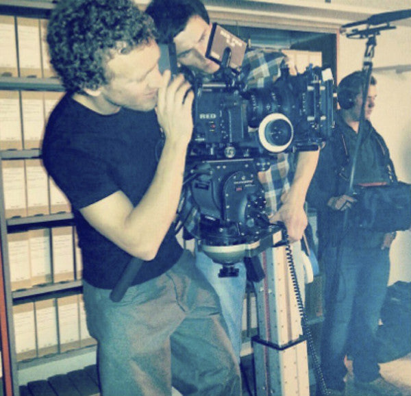 Man in a black shirt looking into a video camera while another man looks on and a sound technician with a microphone stands nearby.