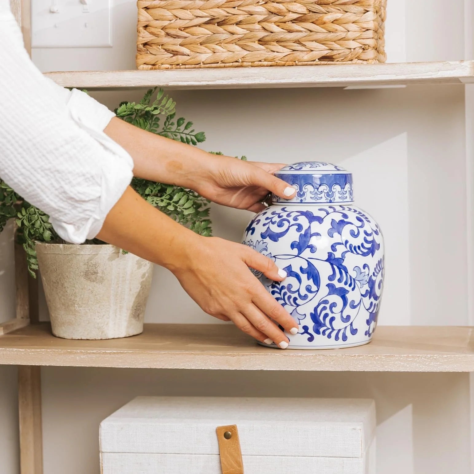 The hands of a woman in a white blouse is placing a blue and white decorative ceramic urn for human ashes on display in the home on a shelf.