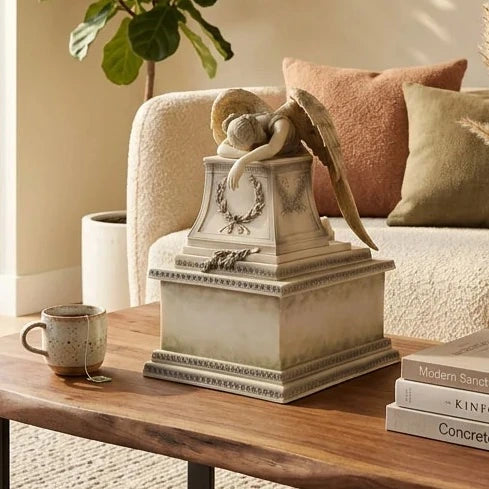 A decorative antique look angel urn or keepsake box with a winged figure draped over the lid, displayed on a wooden table in a living room setting with a sofa, pillows, mug, and books.