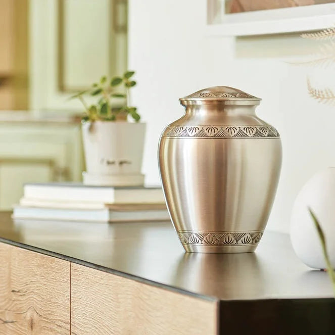 A brushed brass metal urn with decorative engraved bands and a matching lid, displayed on a dark countertop in a softly lit home.