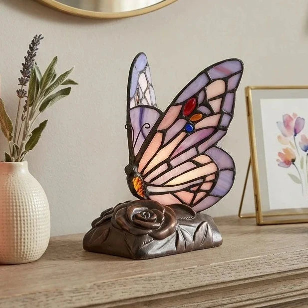 A stained-glass butterfly urn lamp with purple, pink, and blue mounted on a dark bronze rose-shaped base, displayed  in a home on a wooden shelf.