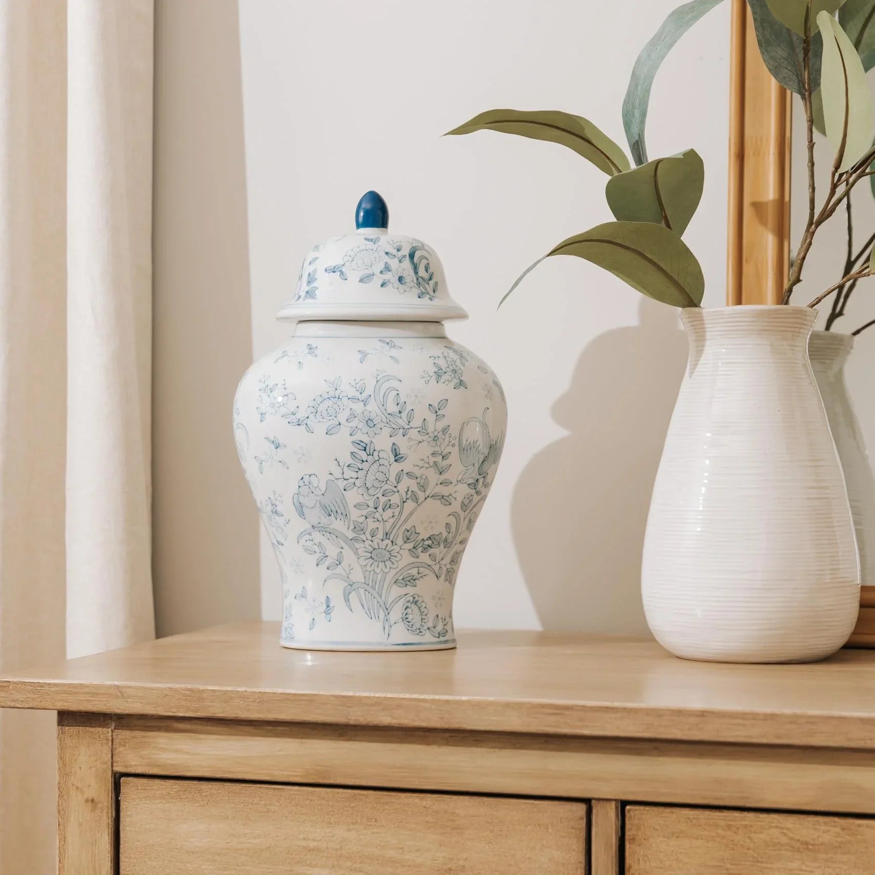 A white ceramic ginger cremation urn for human ashes with a blue floral toile pattern and a blue finial lid, displayed in a home on a light wood cabinet next to a white vase with green leaves.