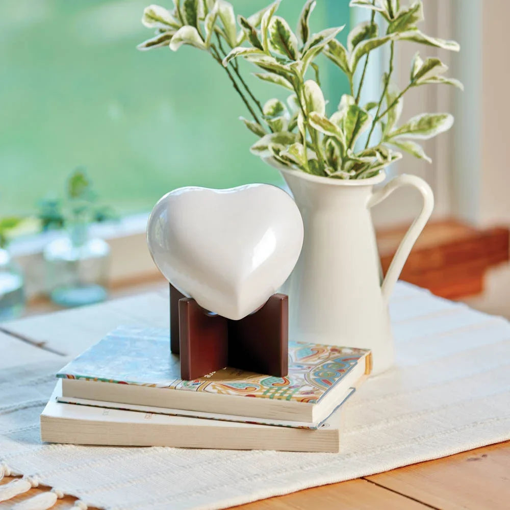 A glossy white ceramic heart urn displayed on a dark brown wooden stand, styled on top of two books next to a white pitcher filled with green and white stems.