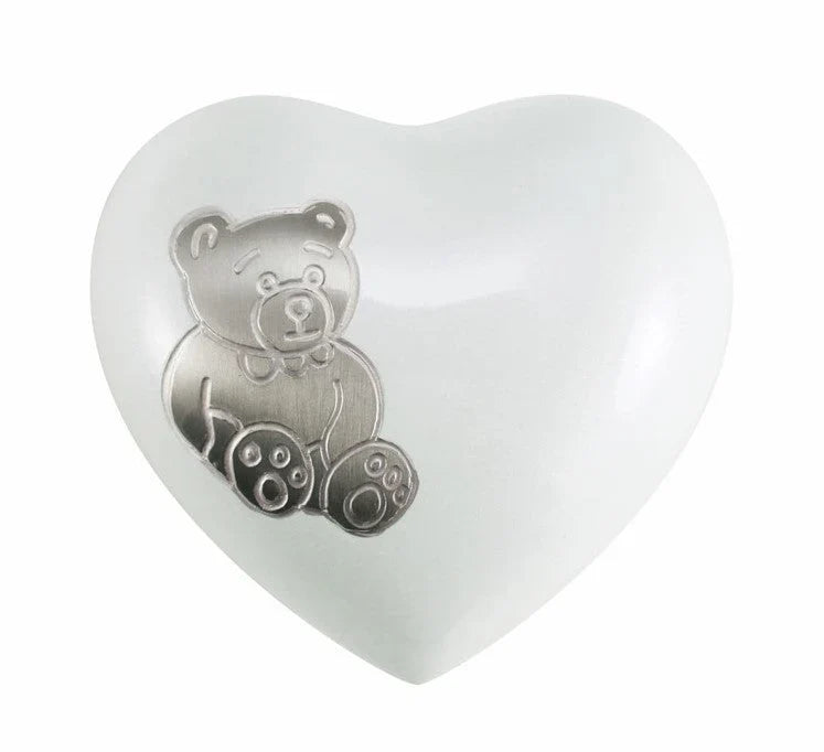 A white heart-shaped cremation urn for  an infant featuring a raised silver teddy bear design on the front, set against a plain white background.