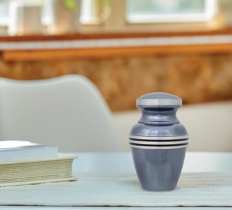 A small urn keepsake cremation container with a metallic blue-gray finish and silver bands, standing on a table in a softly blurred indoor setting.