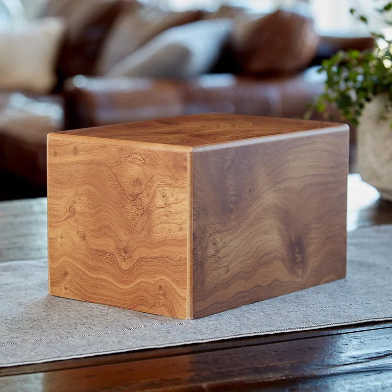 A square wooden cremation urn with a rich unique wood grain finish, shown in a cozy living room setting.