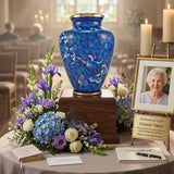 Blue cloisonne cremation urn with a butterfly pattern and floral design, sitting on table at a memorial celebration of life service, on a wooden pedestal with a flower arrangement and photograph of grandma.