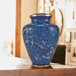 Blue cloisonne cremation urn with a butterfly pattern and floral design, sitting on a wooden table in a warm toned room with a blurry background.