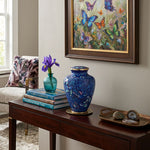 Blue cloisonne cremation urn with a butterfly pattern and floral design, sitting on a welcoming console table styled with books and bluttefly artwork above.