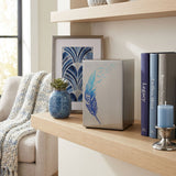 A metal cremation urn featuring a blue feather design that transcends into birds taking flight in a home decor setting.