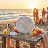Round white ceramic cremation urn with soft, polished wave pattern in a beach celebration of life setting.
