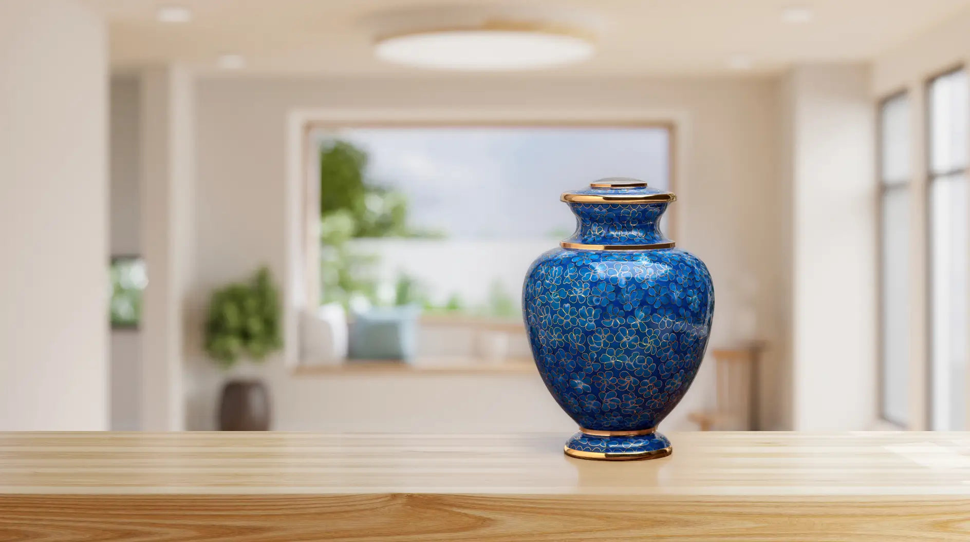 A blue cloisonne hand crafted metal urn with a floral pattern and gold trim, sitting on a light wooden surface with a softly blurred indoor background.