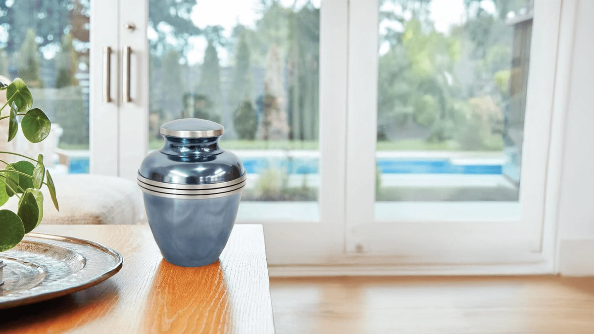 A metallic blue urn with silver trim sits on a wooden table in a bright room with large windows overlooking a pool and garden.