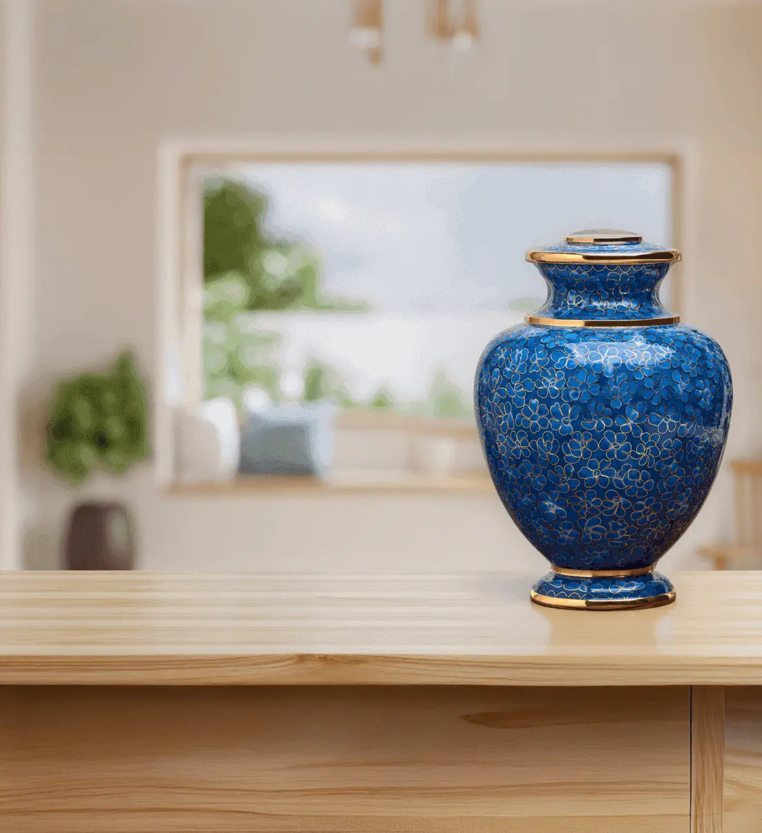 A decorative blue cremation urn with gold cloisonne detailing and a matching lid, displayed on a light wood tabletop in a softly blurred modern interior setting.