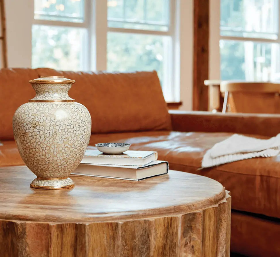A cloisonne urn for ashes with a beige floral patterned on diplsay on a rustic round wood coffee table in a cozy living room setting.