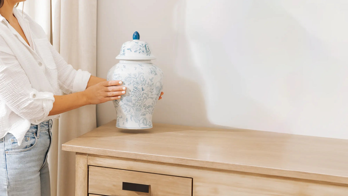 A woman in a white blouse and jeans holds a blue and white patterned ceramic ginger jar cremation urn with a lid, displayed in the home on a light wood dresser against a white wall.