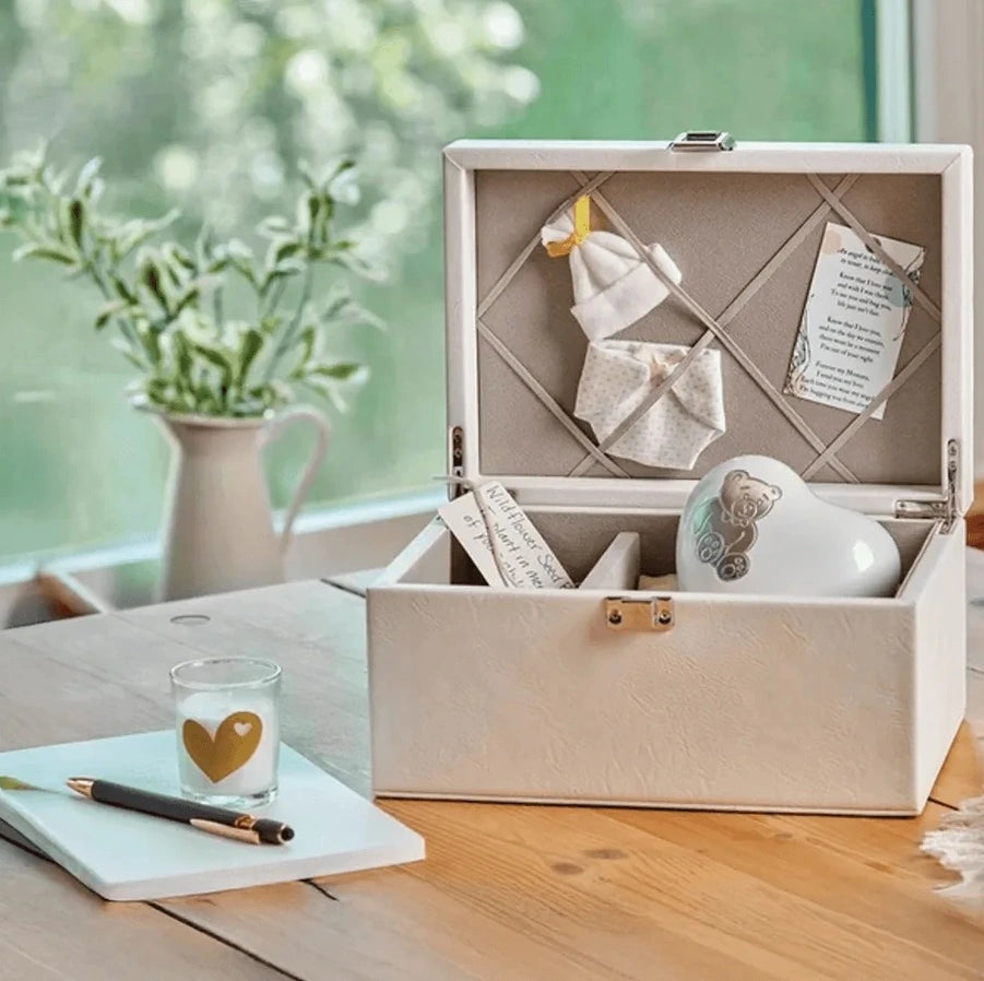 An open cream-colored keepsake memory box on a wooden table, containing a white ceramic teddy bear infant urn.