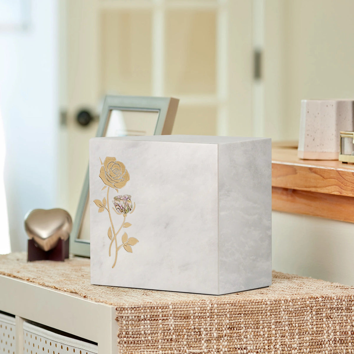 A square white marble cremation urn with a gold rose etched on the front, displayed on a woven surface in home.