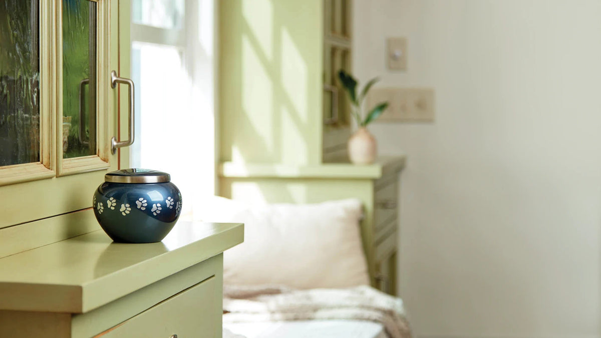 A dark blue pet urn with silver paw prints and a silver banded lid sits on a light green dresser in a softly lit bedroom.