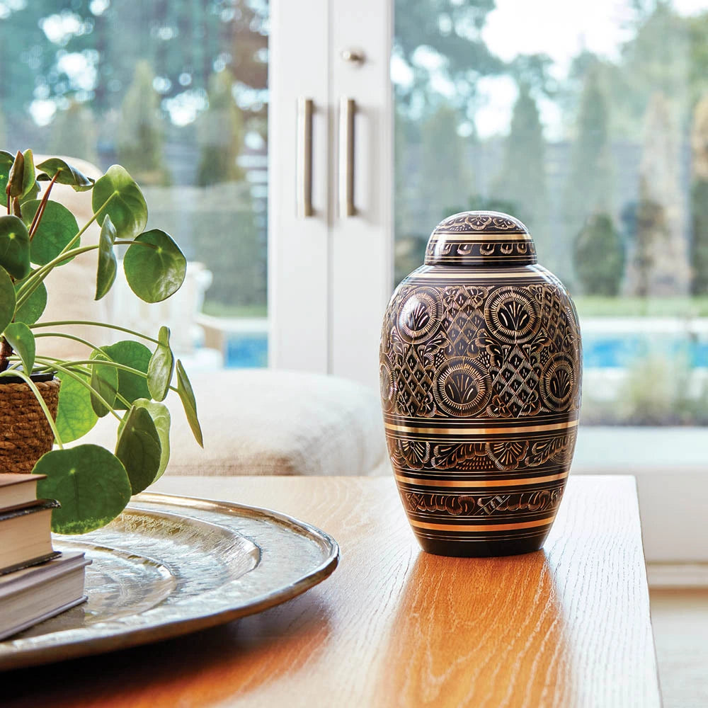 Large decorative black urn with gold detail sits on a table with a plant.