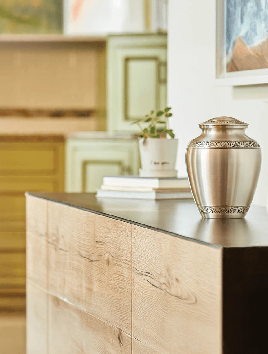 A brushed silver metal urn with a decorative embossed border sits on a dark countertop next to a light wood credenza in a modern, softly blurred living space.