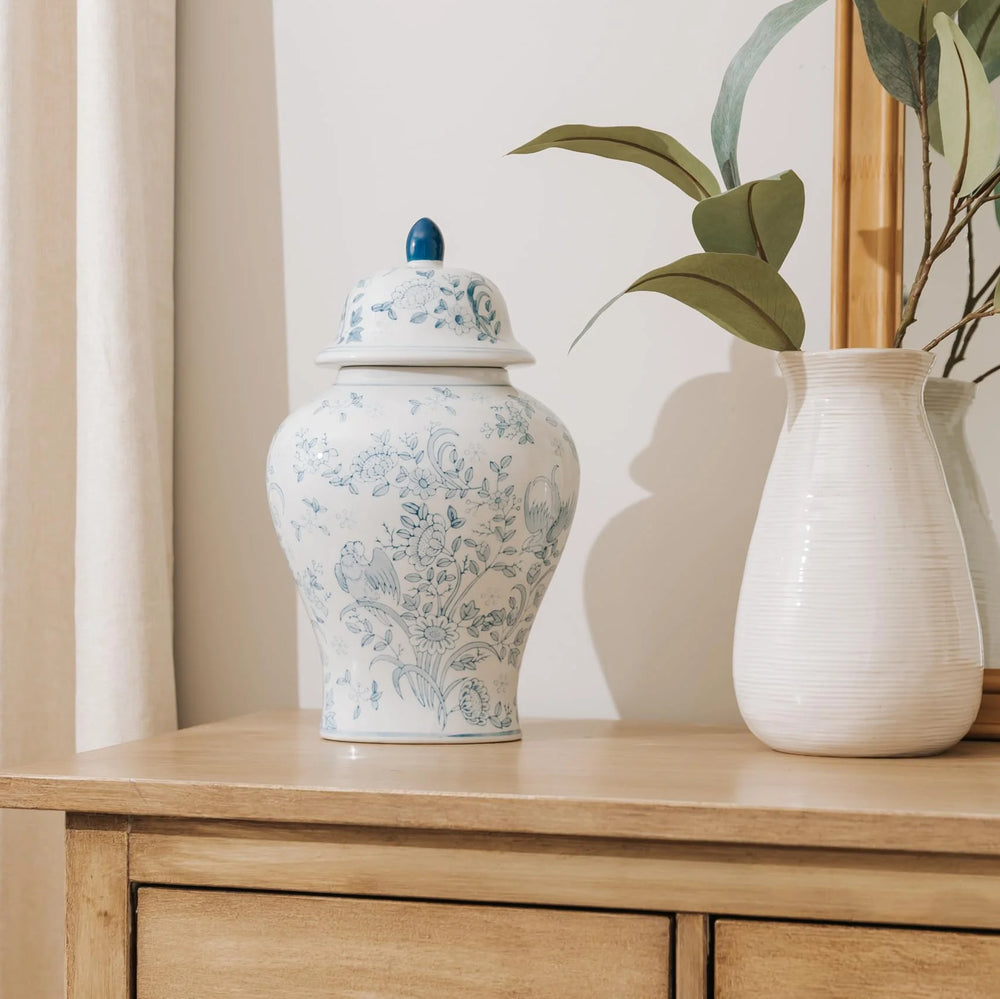 A white ceramic ginger cremation urn for human ashes with a blue floral toile pattern and a blue finial lid, displayed in a home on a light wood cabinet next to a white vase with green leaves.