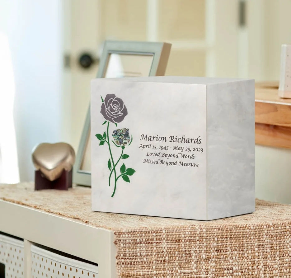 A personalized white marble urn in a box shape with a purple and green rose etched, displayed on a woven surface in a softly lit indoor setting.