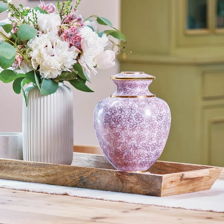 A decorative urn for mom with a pink floral pattern and gold trim sits on a rustic wooden tray next to a white vase holding a bouquet of white and pink flowers, on display in a softly lit home.