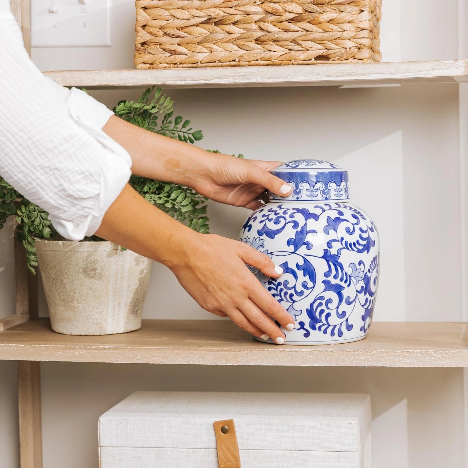 The hands of a woman in a white blouse is placing a blue and white decorative ceramic urn for human ashes on display in the home on a shelf.