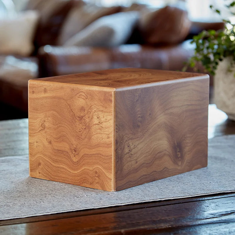 A square wooden cremation urn with a rich unique wood grain finish, shown in a cozy living room setting.