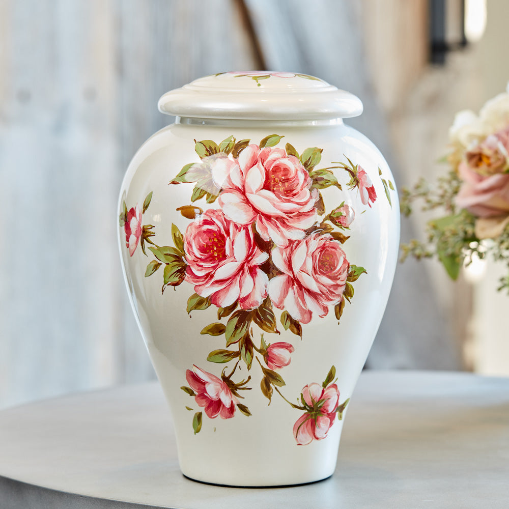 White ceramic porcelain urn with delicate pink roses sitting on a table in a brightly lit room. 